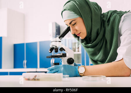 concentrated female muslim scientist looking through microscope during experiment in chemical laboratory Stock Photo