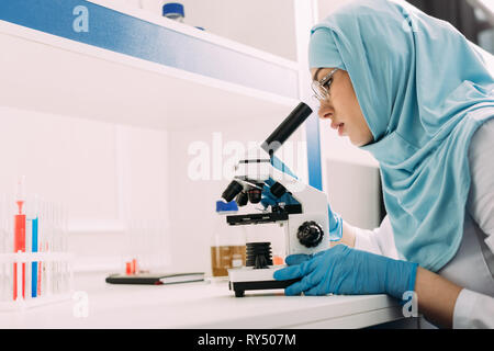female muslim scientist looking through microscope during experiment in chemical laboratory with copy space Stock Photo