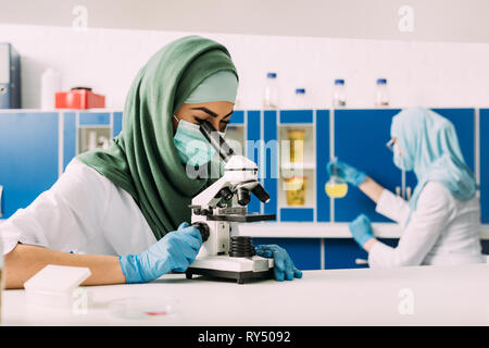 female muslim scientist looking through microscope during experiment with colleague working on background in chemical laboratory Stock Photo