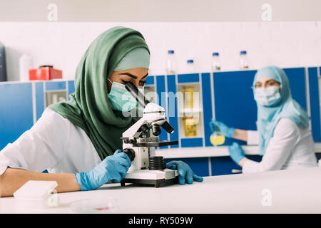 female muslim scientist looking through microscope during experiment with colleague working on background in chemical laboratory Stock Photo