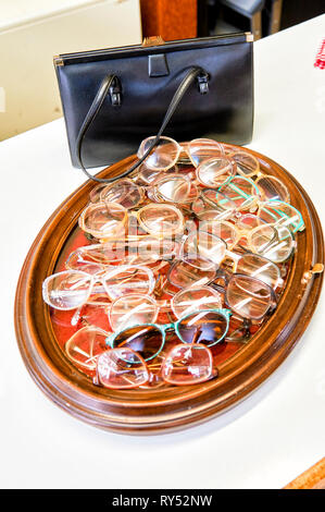 Auf einem Bilderrahmen liegen viele verschiedene Brillengestelle in einem Gebrauchtwarenlgeschaeft. Dahinter steht eine schwarze Damenledertasche Stock Photo