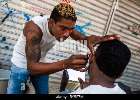 A young Colombian man shaves a friend’s beard with a razor blade in the market of Bazurto, Cartgena, Colombia. Stock Photo