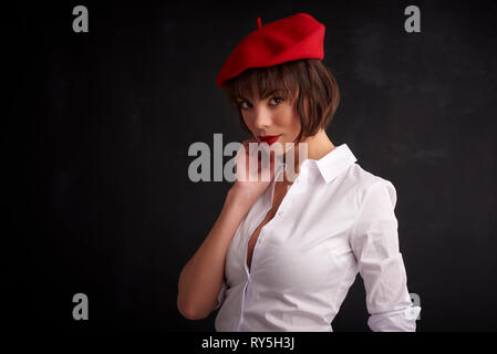 Portrait shot of beautiful young woman wearing red beret and white shirt while posing at dark background. Stock Photo