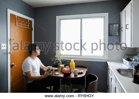 Thoughtful young Latinx man looking out window in morning kitchen
