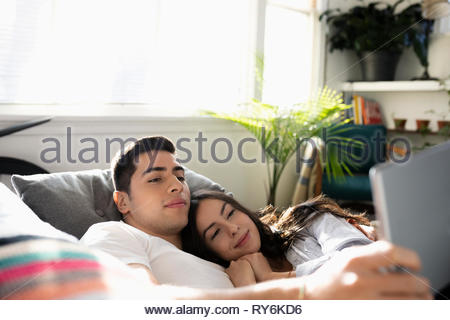 Serene young Latinx couple cuddling on sofa, using digital tablet