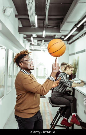Smiling positive African American guy playing with basketball ball in office Stock Photo