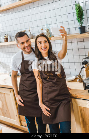 cashiers in aprons standing, smiling and taking selfie in coffee house Stock Photo