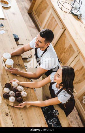 cashiers in brown aprons offering cupcakes and coffee in paper cups behind wooden bar counter in coffee house Stock Photo