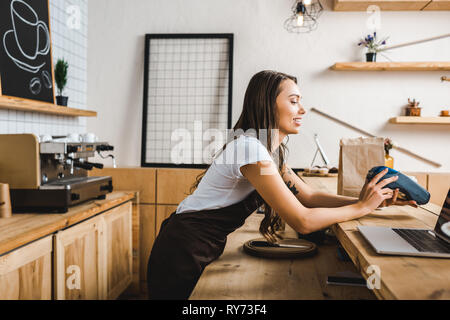 attractive cashier standing in brown apron and holding terminal behind bar counter in coffee house Stock Photo