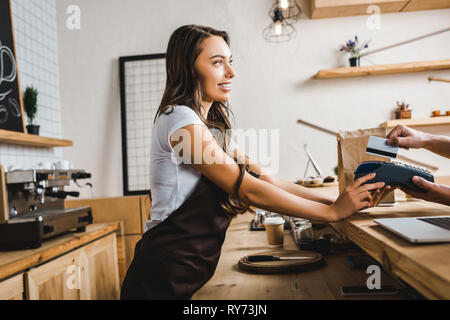 attractive cashier standing in brown apron and holding terminal wile man paying with credit card in coffee house Stock Photo