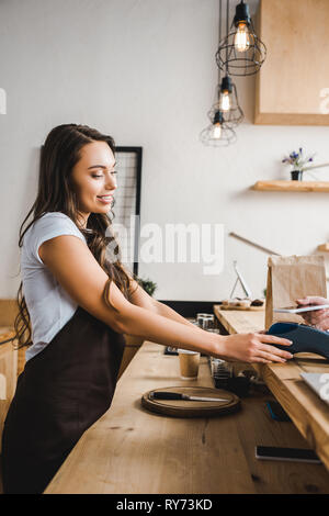 attractive cashier standing in brown apron and holding terminal wile man paying with smartphone in coffee house Stock Photo