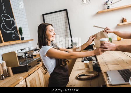 attractive cashier in brown apron giving paper cup and bag to man in coffee house Stock Photo