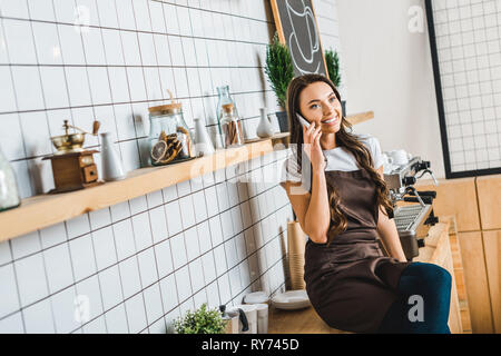 attractive cashier in brown apron talking on smartphone near coffee machine and sitting on bar counter in coffee house Stock Photo