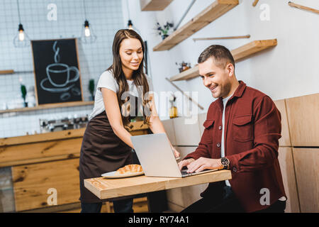 attractive waitress standing near freelancer sitting at table, looking to laptop and smiling in coffee house Stock Photo
