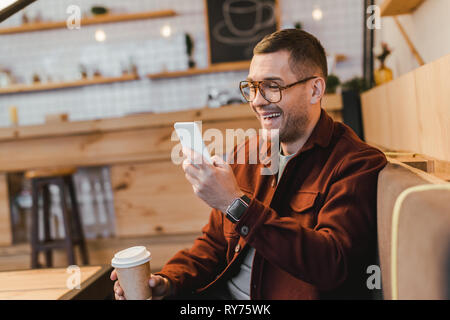 handsome man in burgundy shirt sitting on couch, holding paper cup, looking to smartphone and laughing in coffee house Stock Photo