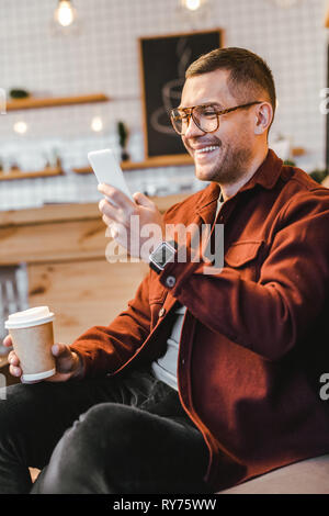 man in burgundy shirt sitting on couch, holding paper cup, looking to smartphone and laughing in coffee house Stock Photo