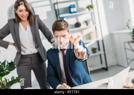 selective focus of paper plane in hand of businessman sitting in office with female coworker on background Stock Photo