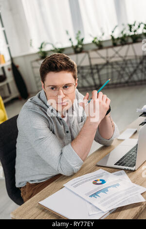 handsome man in glasses holding pencil and sitting near charts and graphs at home Stock Photo
