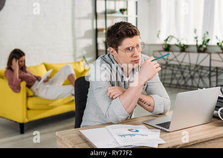 pensive man holding pencil and sitting near laptop near girlfriend on sofa Stock Photo