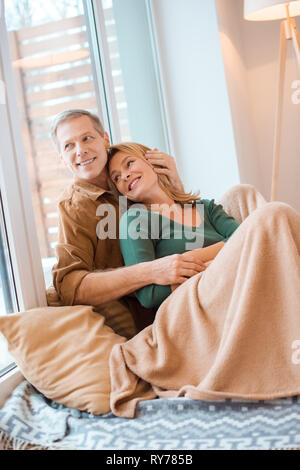 smiling couple hugging while sitting on floor by large window at new home Stock Photo