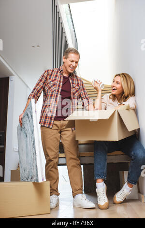 smiling man holding picture while standing near wife sitting on stairs with carton box Stock Photo