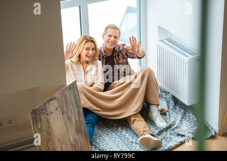 happy couple waving hands while sitting on floor by large window at new home Stock Photo