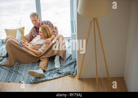 happy couple using laptop while resting on floor by large window at new home Stock Photo