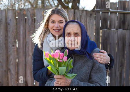 Young woman giving a bouquet of pink tulips to her elderly grandmother  on Women's day, 8th of March. Love, care, gift, togetherness Stock Photo