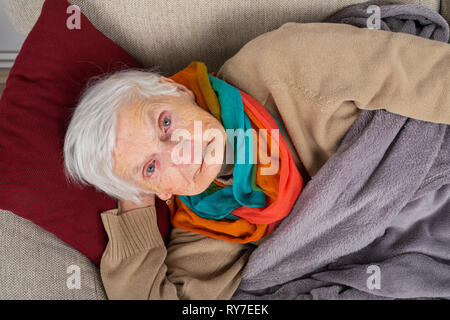 Close up picture of a sick old lady lying on the couch wrapped in a blanket, wearing colorful scarf Stock Photo