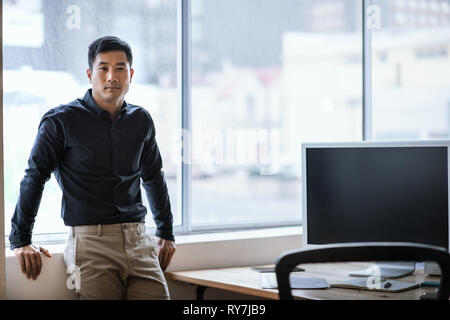 Confident young Asian businessman standing by windows in an office Stock Photo