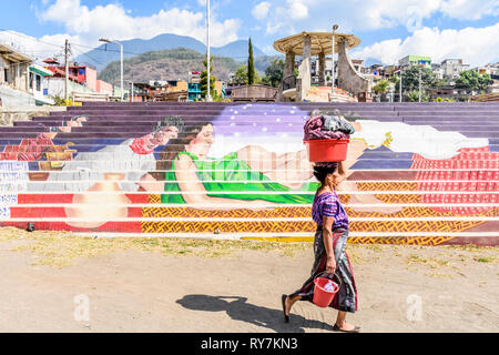 Santiago Atitlan, Lake Atitlan, Guatemala - March 8, 2019: Traditionally dressed Maya woman walks past colorful mural of childbirth in lakeside town Stock Photo