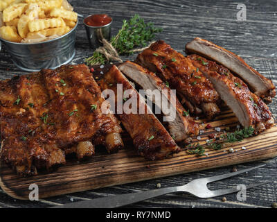 cutting grilled pork ribs with sauce on a board, french fries, spices, wooden background Stock Photo