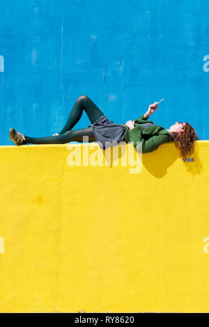 Side view of businesswoman using smart phone while lying on yellow wall during sunny day Stock Photo