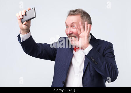 Senior man looking at screen of smart phone, waving hello as having video chat Stock Photo