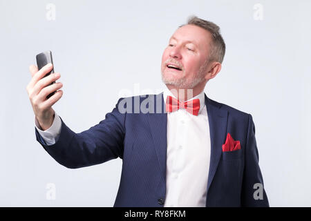 Senior man in blue suit looking at screen of smart phone, smiling nicely Stock Photo