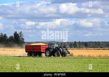 Farm tractor pulls agricultural trailer load of harvested grain along small rural road in Finnish countryside under blue sky and white clouds. Stock Photo