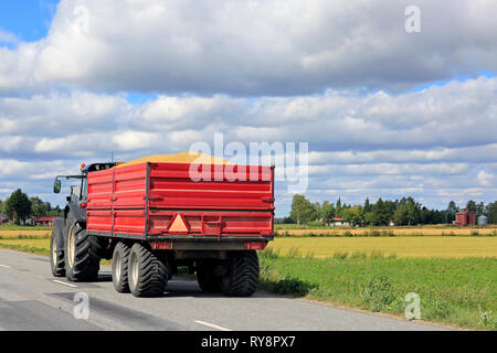 Tractor pulls red agricultural trailer with a full load of harvested grain along country road on a sunny day in autumn harvest time. Stock Photo