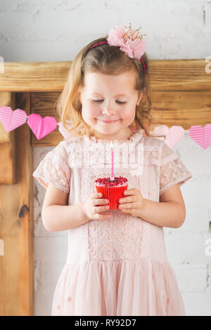 Little girl in pastel pink dress and crown headband holding red velvet cupcake with lit up candle, solid wood mantelpiece background, selective focus Stock Photo