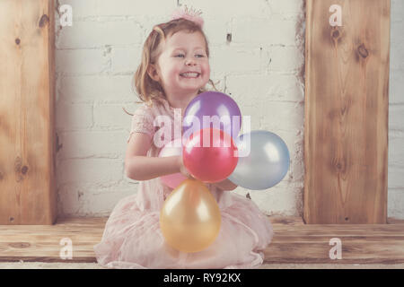 Little girl in pastel pink dress and crown headband holding pastel colored and vibrant baloons, solid wood mantelpiece background, toned, selective focus Stock Photo