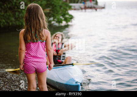 Rear view of granddaughter standing on lakeshore while grandmother sitting in kayak Stock Photo