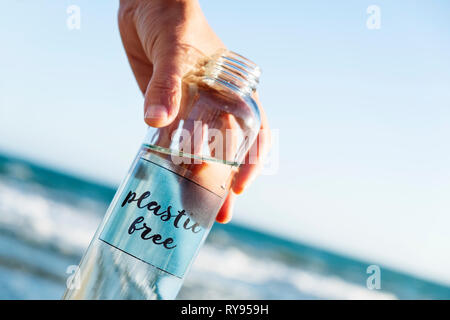 closeup of a caucasian man holding a glass reusable water bottle with the text plastic free written in it, on the beach, with the ocean in the backgro Stock Photo