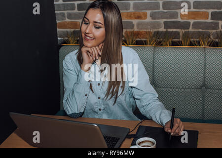 Female graphic designer working on laptop computer while using graphic tablet at desk in cafe Stock Photo