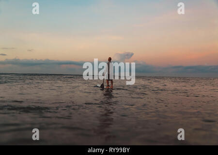 Back view of silhouette of male with paddle on surf board between water of sea and sky in evening on Bali, Indonesia Stock Photo