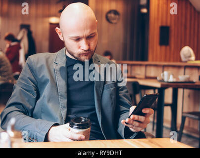 Adult bald gloomy man drinking coffee from paper cup and using mobile phone at cafe Stock Photo
