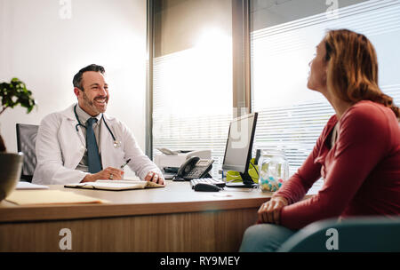 Doctor talking with female patient sitting across the desk in clinic. Mature male doctor in white medical coat consulting his patient. Stock Photo