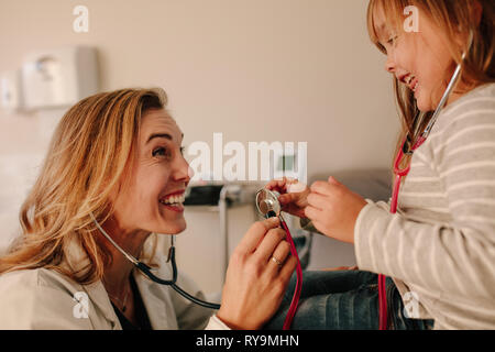 Young pediatrician playing with her girl patient. Friendly doctor and young girl on medical appointment, both playing with stethoscope. Stock Photo