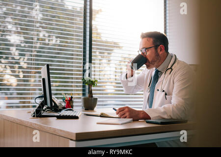 Mature male doctor drinking coffee at his desk. Doctor working at his office desk and drinking coffee. Stock Photo