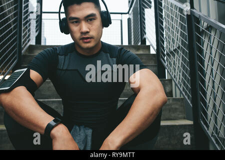 Portrait of asian man relaxing after workout sitting outdoors on stairs. Male athlete listening to music using wireless headphones while relaxing afte Stock Photo