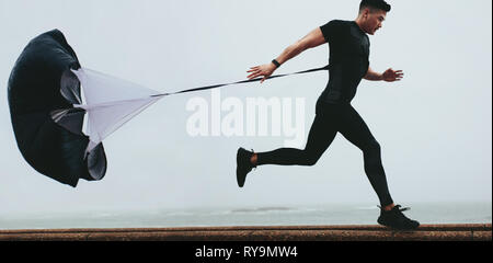 Strong man doing workout using resistance parachute outdoors. Young asian man in black sports clothing running with parachute. Stock Photo