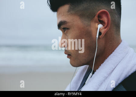 Close up of young asian man with earphone and towel around his neck. Male fitness model listen music with earphones while taking rest from exercising. Stock Photo